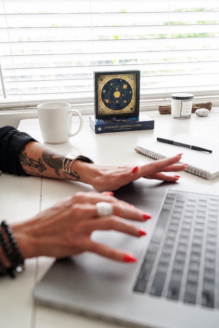 Hands at a laptop with astrology reading cards and a notebook on the desk